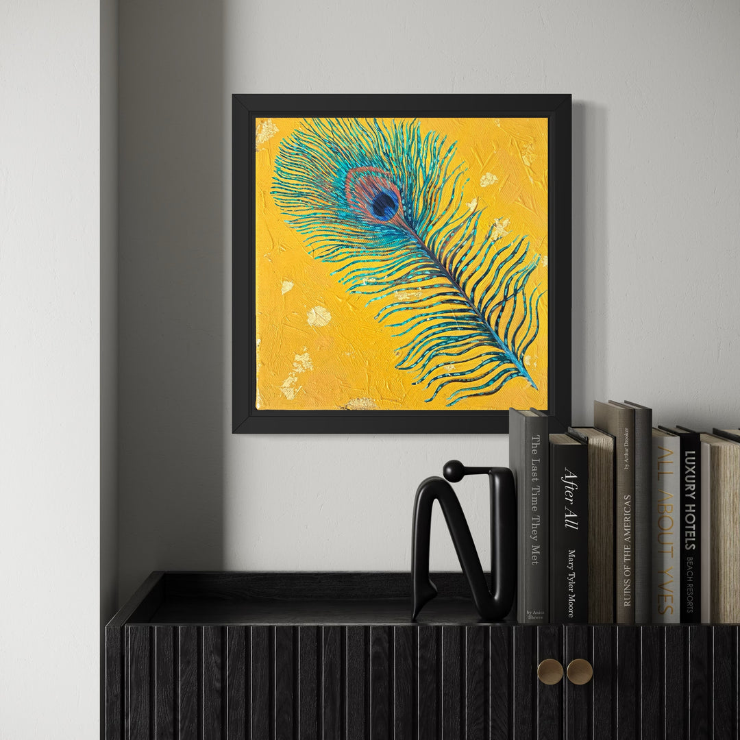 Framed artwork of a peacock feather on a yellow background, displayed on a shelf with books.