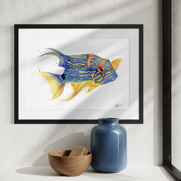 Framed artwork of a colorful fish on a white wall with a blue vase and wooden bowl below.