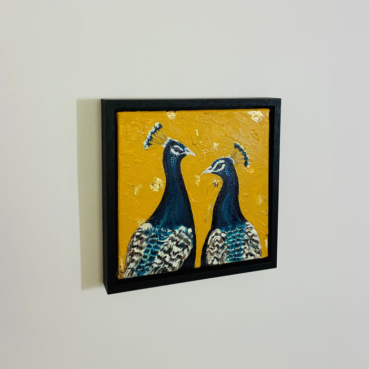 Two framed artworks of peacocks on a white wall.