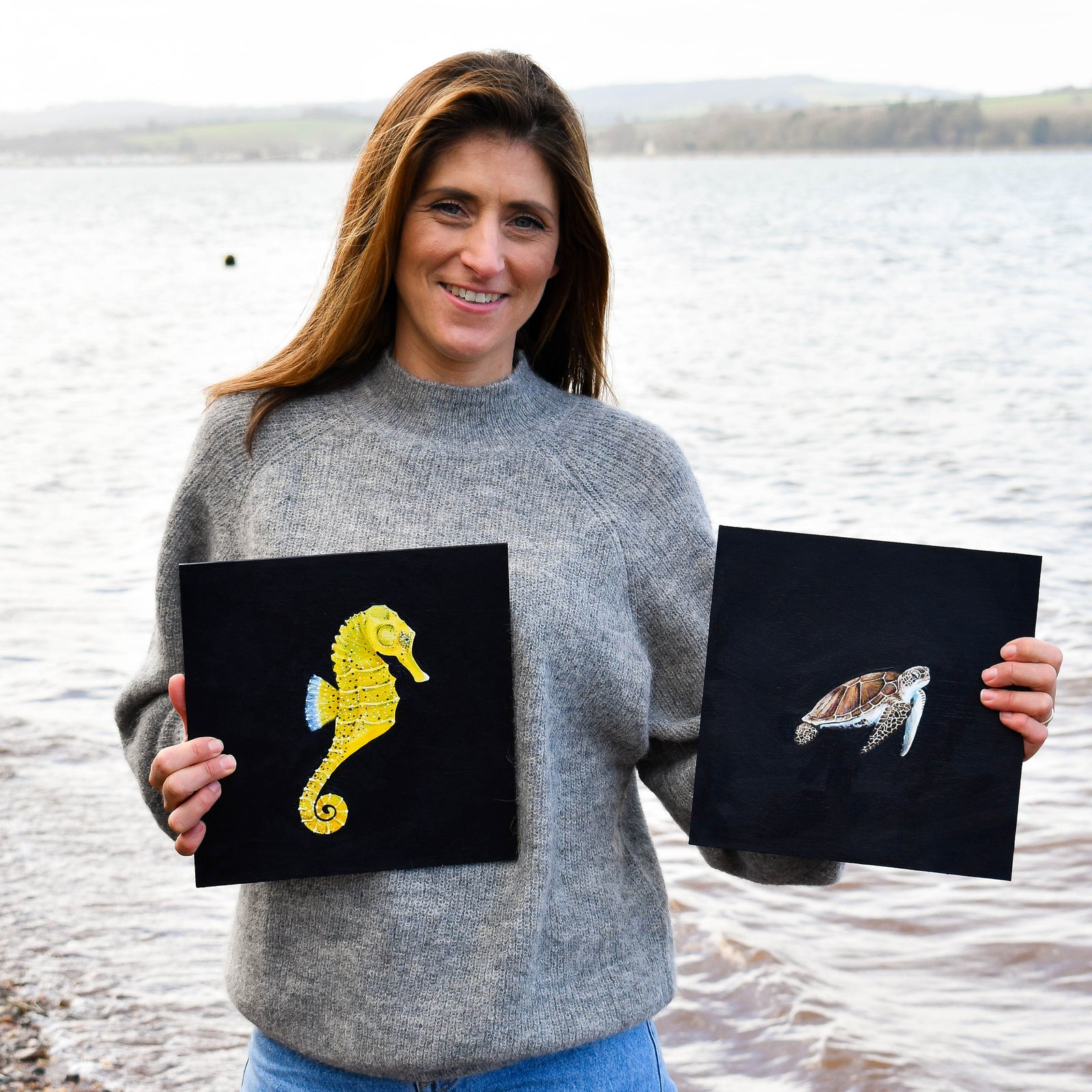 Woman holding two black cards with animal illustrations by a body of water.