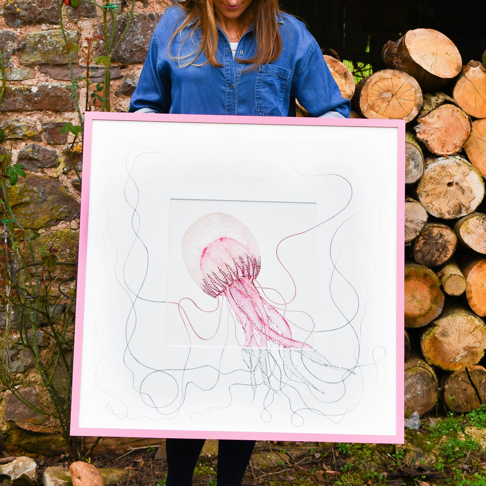 Person holding a framed artwork of a jellyfish in front of a brick wall and stacked logs.